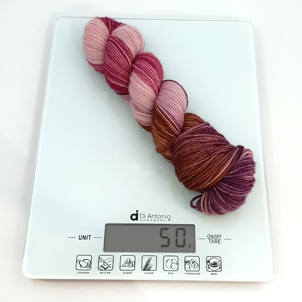 Hand-dyed yarn skein in variegated pink-purple-brown tones on weigh scales displaying 50g